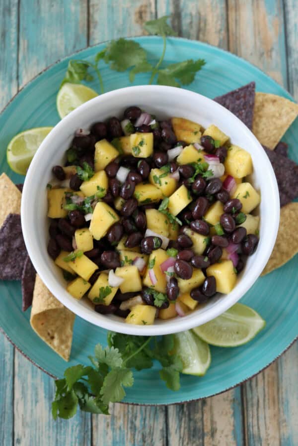 A bowl of black bean and mango salsa with chopped onions and cilantro, served on a blue plate with tortilla chips, lime wedges, and cilantro sprigs on a rustic wooden table.
