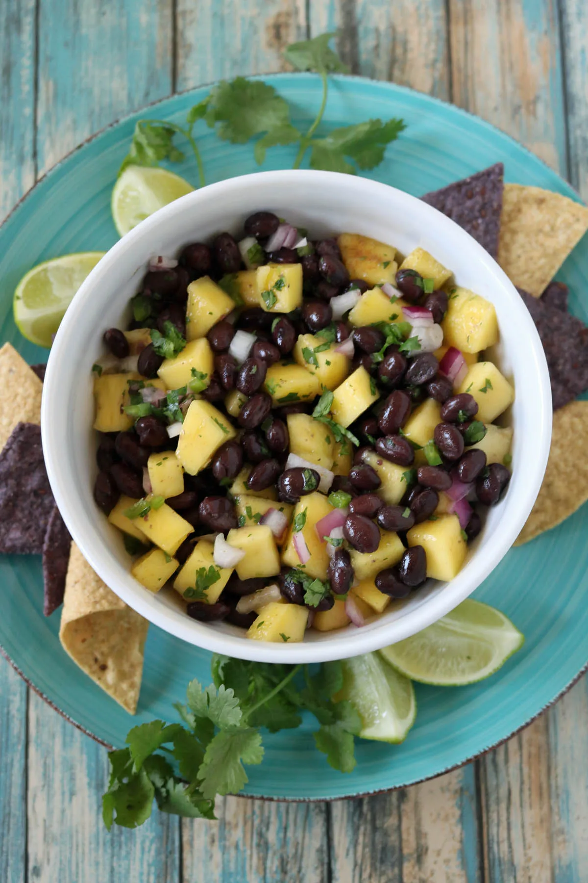 A bowl of black bean and mango salsa with chopped onions and cilantro, served on a blue plate with tortilla chips, lime wedges, and cilantro sprigs on a rustic wooden table.