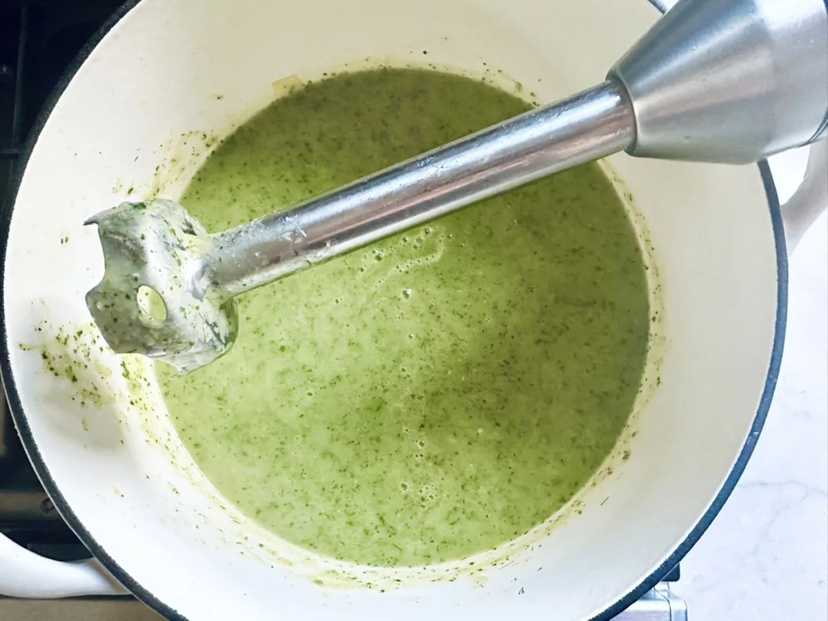 A hand blender is blending zucchini soup with dill and Greek yogurt in a white pot, creating a smooth consistency. The pot sits on a stovetop, and specks of fresh herbs are visible in the vibrant green soup.