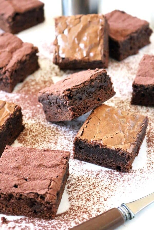 Several thick, fudgy Chocolate Chipotle Brownies with crackled tops are arranged on a white surface dusted with cocoa powder, with a knife nearby.