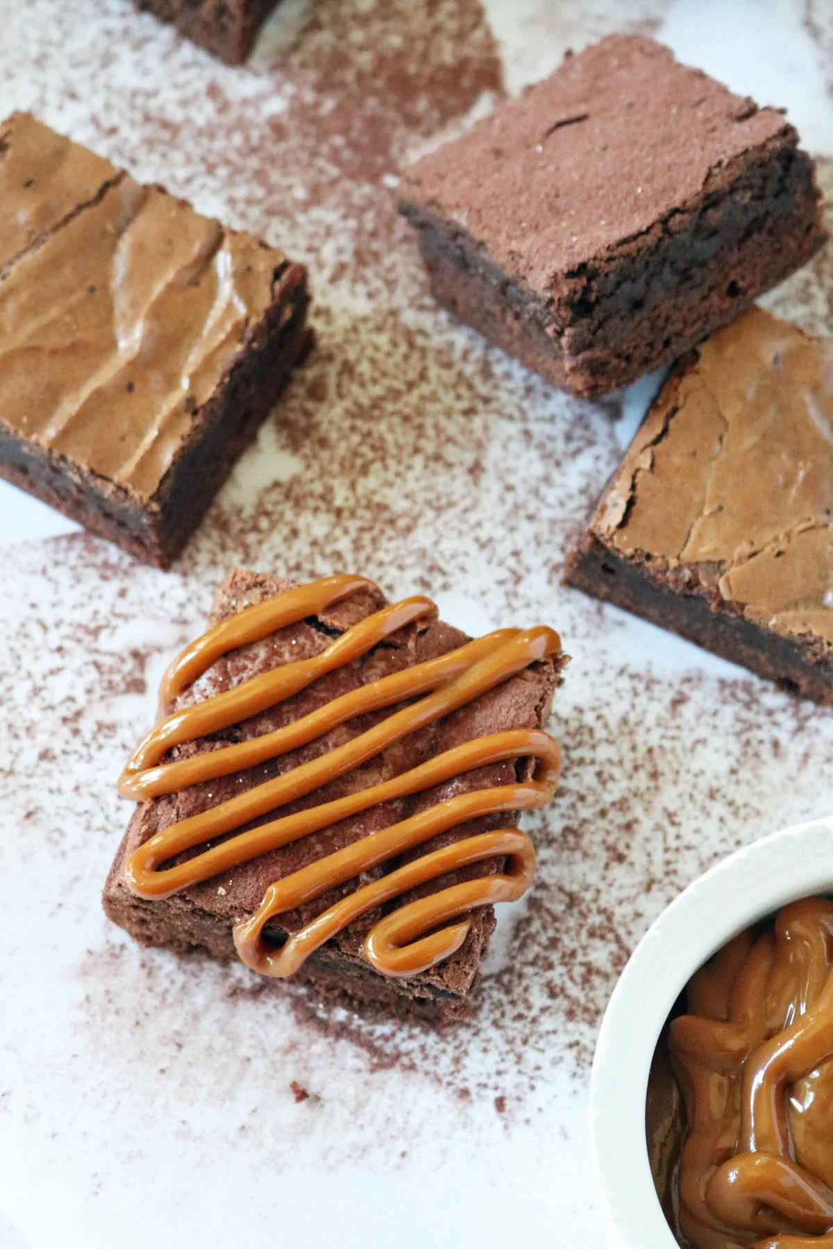 A square Chocolate Chipotle Brownie drizzled with caramel sits on a white surface dusted with cocoa powder, surrounded by more plain brownies. A small bowl of caramel is partially visible in the corner.