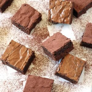 Nine Chocolate Chipotle Brownies are arranged on a white surface dusted with cocoa powder. A knife and a small container of cocoa powder are also visible among the scattered brownies.