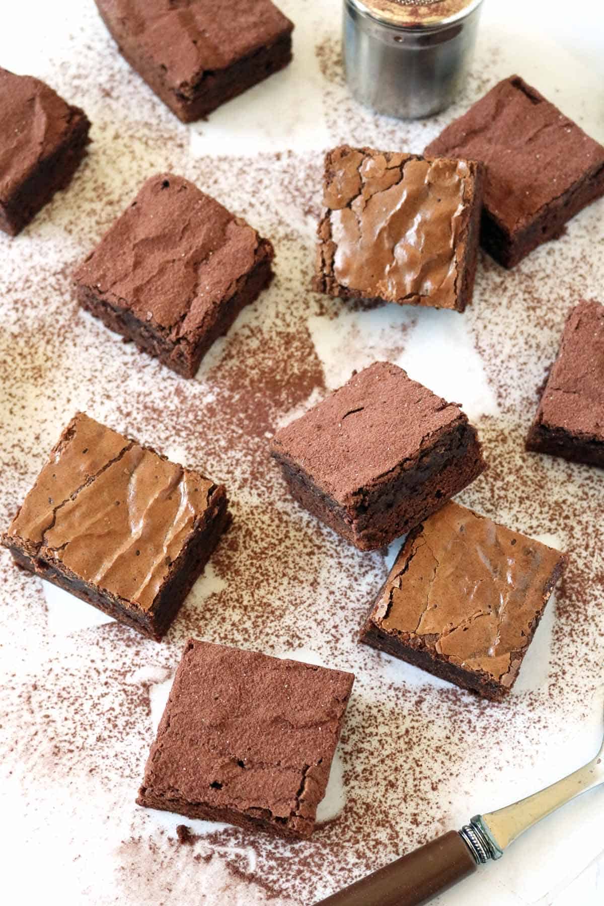 Nine Chocolate Chipotle Brownies are arranged on a white surface dusted with cocoa powder. A knife and a small container of cocoa powder are also visible among the scattered brownies.