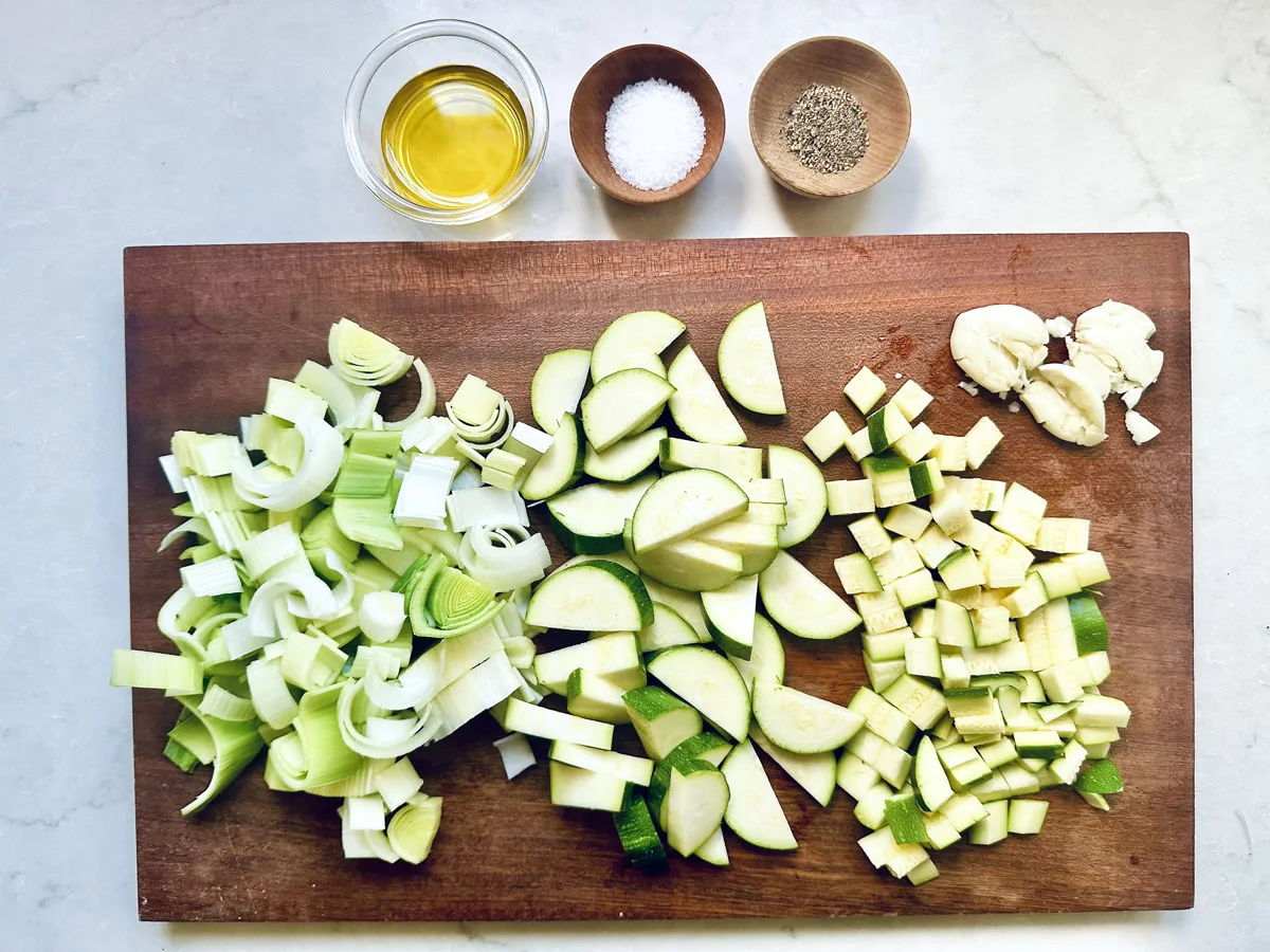A wooden cutting board with chopped leeks, zucchini for Zucchini Soup with Dill and Greek Yogurt, garlic cloves, and small bowls of olive oil, salt, and pepper, all arranged neatly on a light countertop.