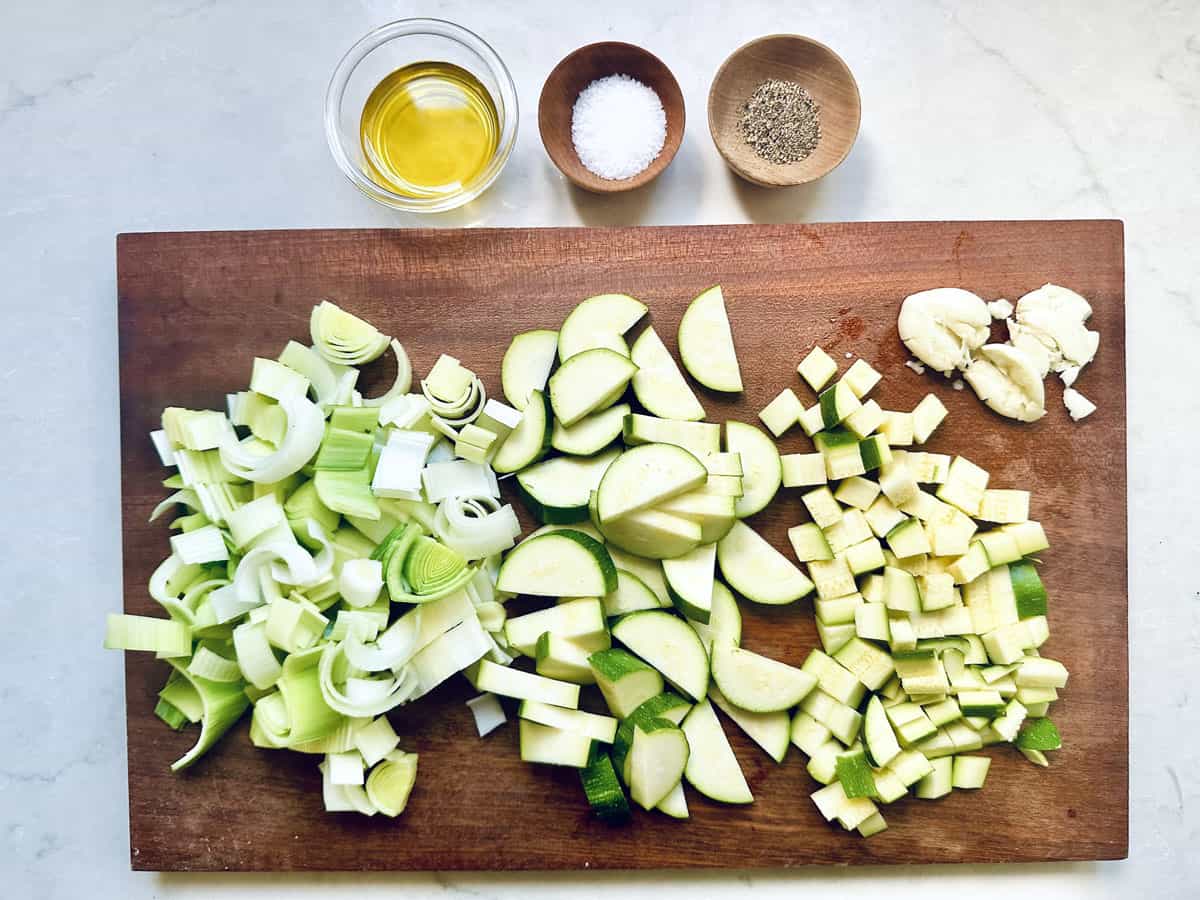 A wooden cutting board with chopped leeks, zucchini for Zucchini Soup with Dill and Greek Yogurt, garlic cloves, and small bowls of olive oil, salt, and pepper, all arranged neatly on a light countertop.