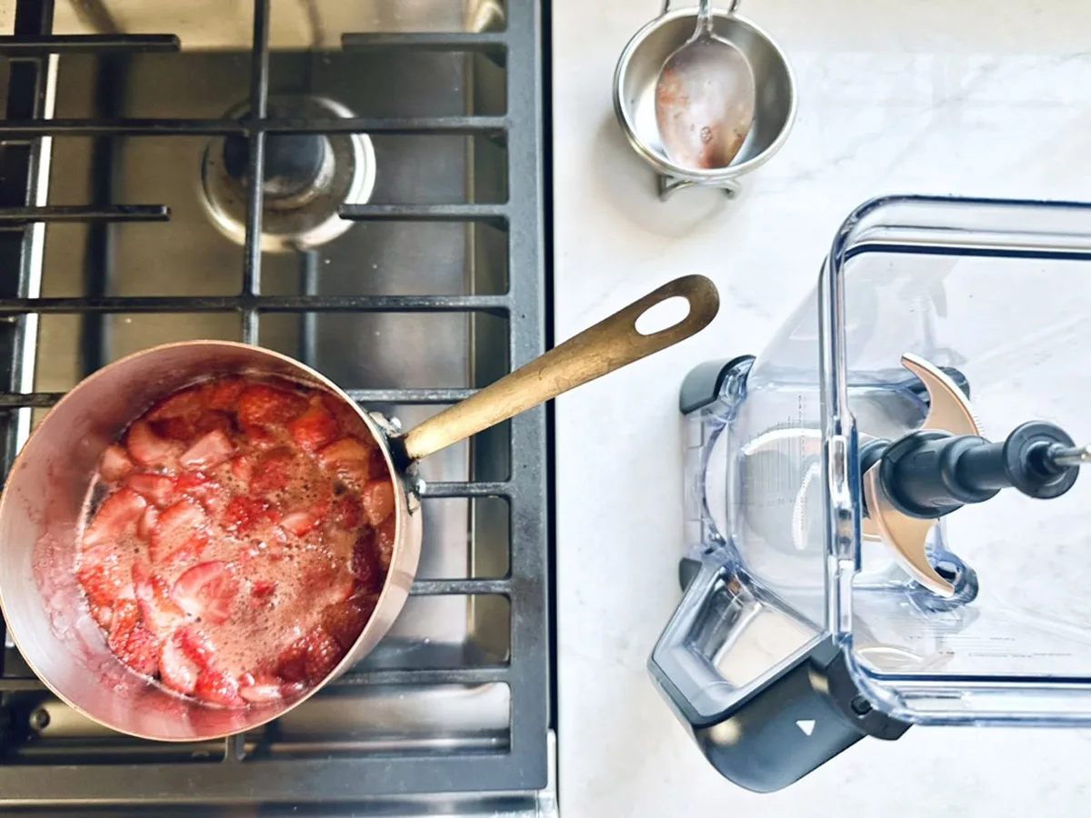 A saucepan with simmering strawberries for Strawberry-Balsamic Ice Cream sits on a stovetop next to a clear blender and a small metal measuring cup on a white countertop.