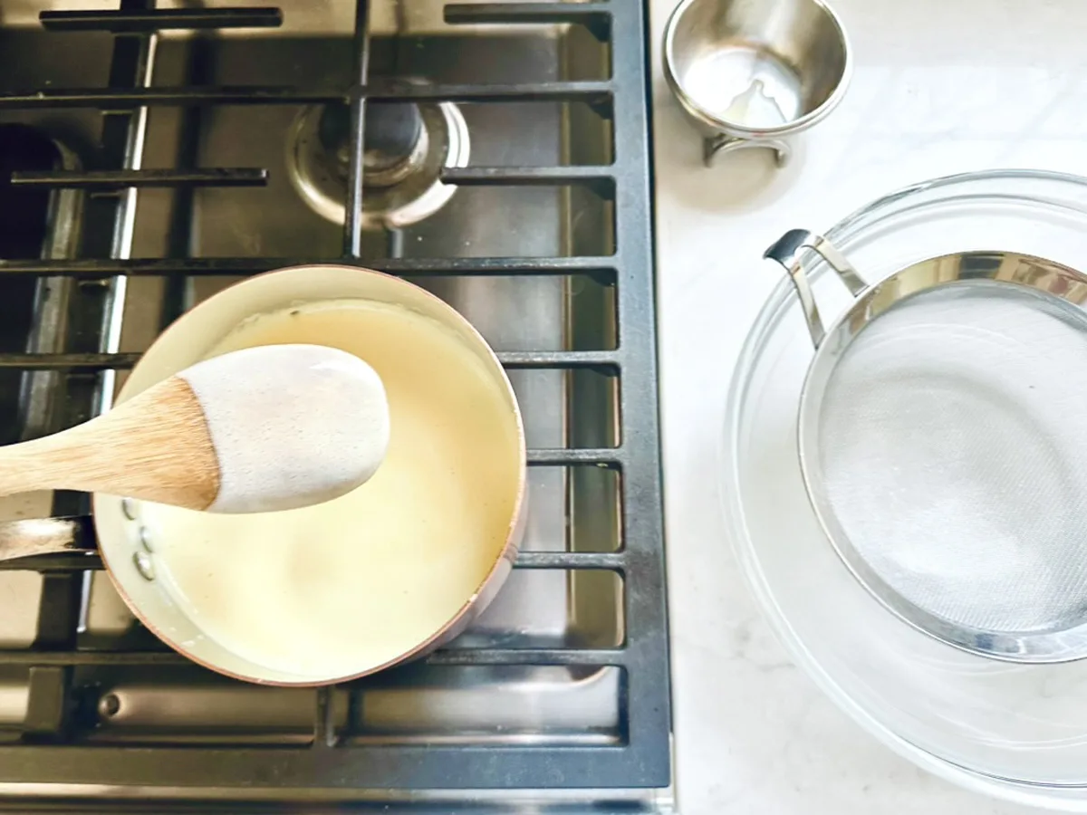 A wooden spoon stirs a creamy Strawberry-Balsamic Ice Cream mixture in a saucepan on the stove, with a metal strainer over a glass bowl and a small metal cup on the white countertop nearby.