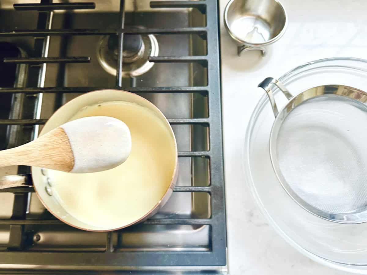 A wooden spoon stirs a creamy Strawberry-Balsamic Ice Cream mixture in a saucepan on the stove, with a metal strainer over a glass bowl and a small metal cup on the white countertop nearby.