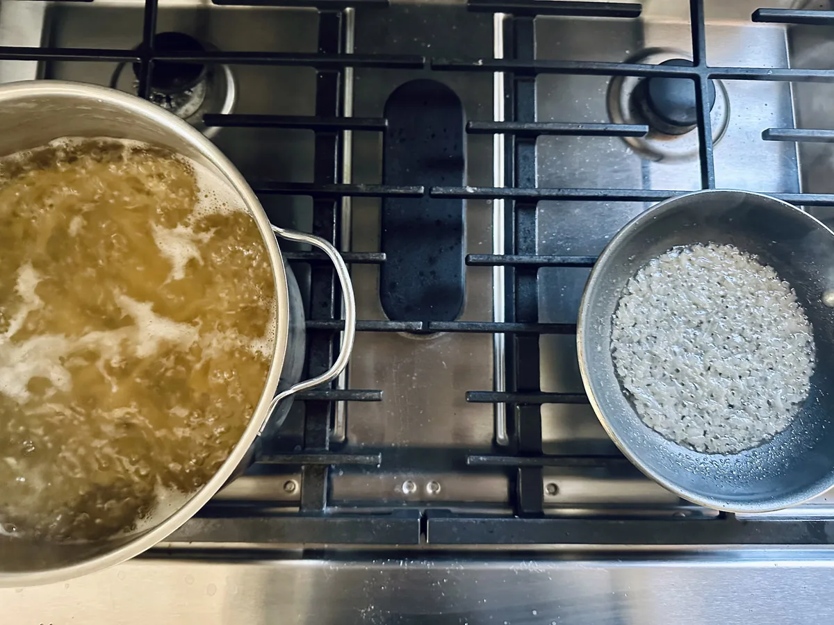A pot of pasta is boiling on the left burner of a stove, while uncooked rice sits in a pan on the right burner. The stove is stainless steel with visible grates and knobs.