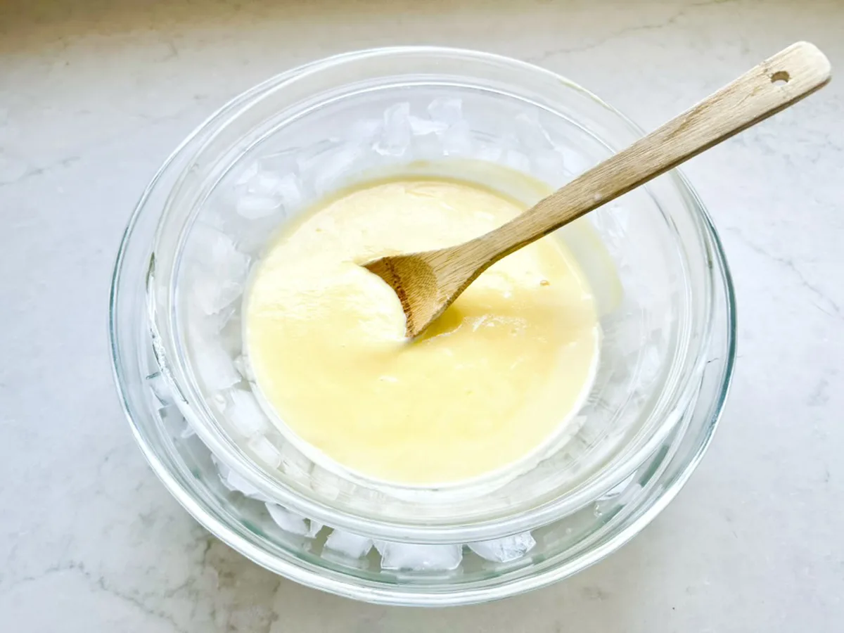 A glass bowl of creamy yellow Strawberry-Balsamic Ice Cream mixture being stirred with a wooden spoon, set inside a larger bowl filled with ice cubes on a white countertop.