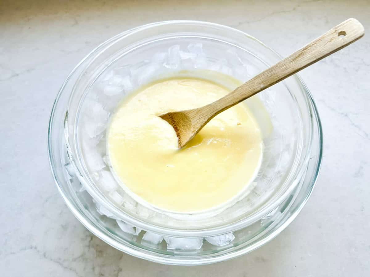 A glass bowl of creamy yellow Strawberry-Balsamic Ice Cream mixture being stirred with a wooden spoon, set inside a larger bowl filled with ice cubes on a white countertop.