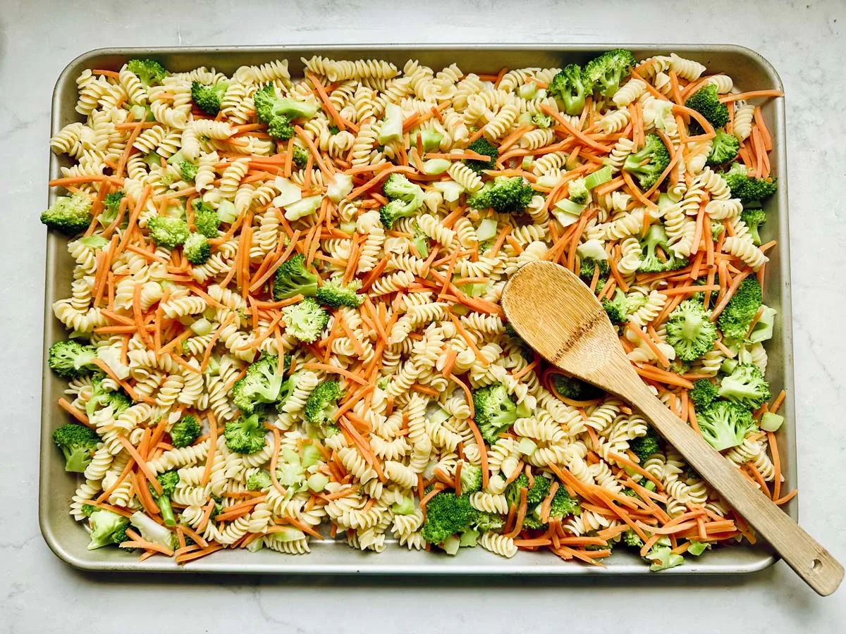A baking sheet filled with uncooked rotini pasta, broccoli florets, and julienned carrots—perfect for a Pasta Salad with Broccoli—rests under a wooden spoon.
