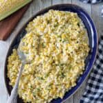 A blue oval platter filled with Corn Salad with Buttermilk and Herbs, topped with chopped herbs, with a serving spoon resting on the dish. Fresh corn on the cob and a checkered cloth sit nearby on a wooden surface.