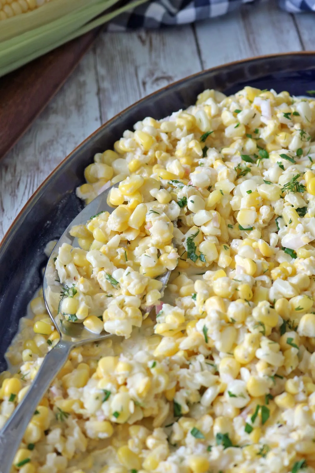 A close-up of a large dish filled with Corn Salad with Buttermilk and Herbs, creamy dressing coating corn kernels, garnished with chopped herbs, and a serving spoon resting in the salad.