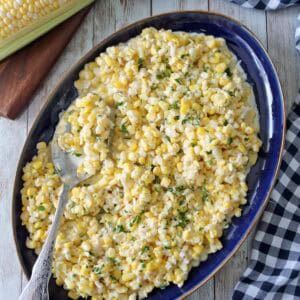 A blue oval platter filled with Corn Salad with Buttermilk and Herbs, topped with chopped herbs, with a serving spoon resting on the dish. Fresh corn on the cob and a checkered cloth sit nearby on a wooden surface.