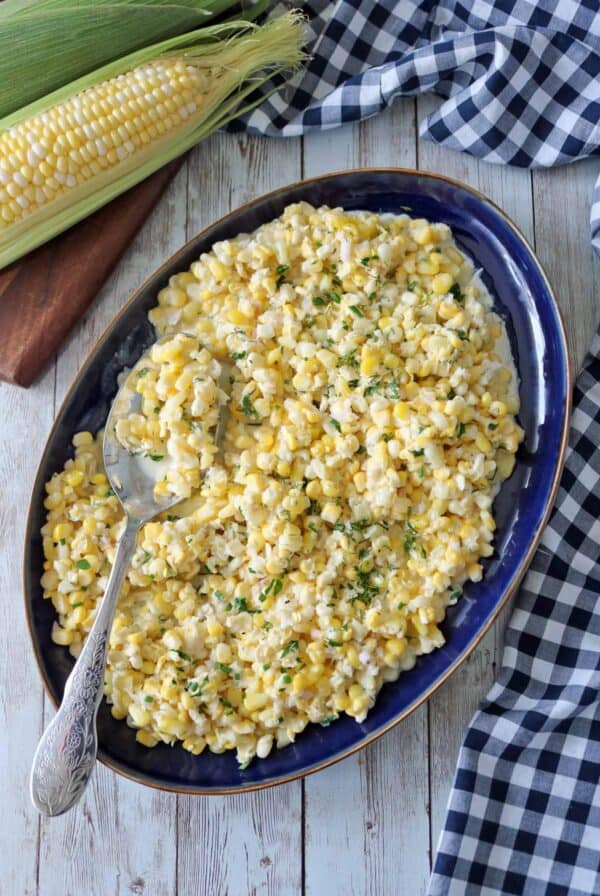 A blue oval platter filled with Corn Salad with Buttermilk and Herbs, topped with chopped herbs, with a serving spoon resting on the dish. Fresh corn on the cob and a checkered cloth sit nearby on a wooden surface.