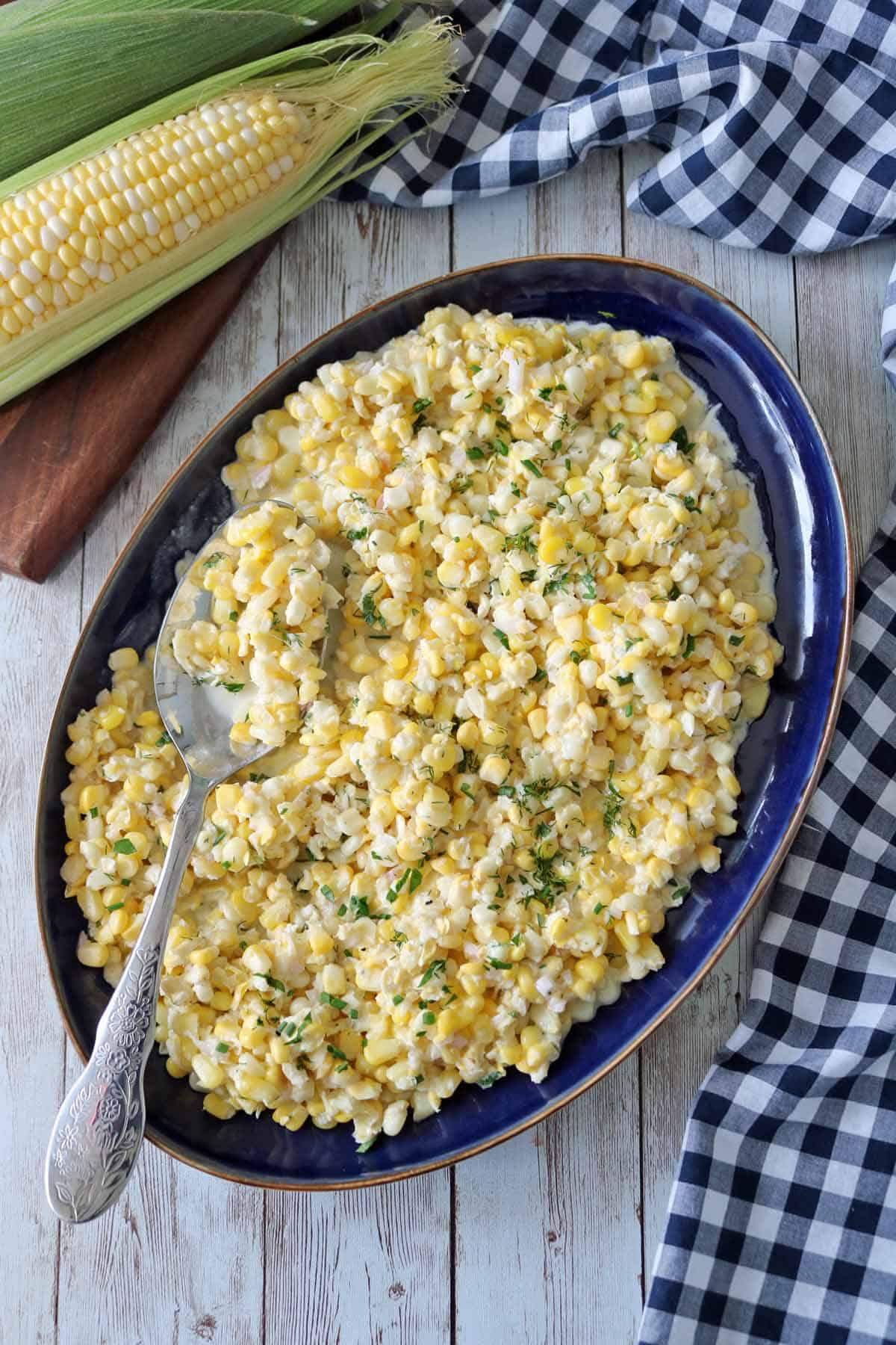 A blue oval platter filled with Corn Salad with Buttermilk and Herbs, topped with chopped herbs, with a serving spoon resting on the dish. Fresh corn on the cob and a checkered cloth sit nearby on a wooden surface.