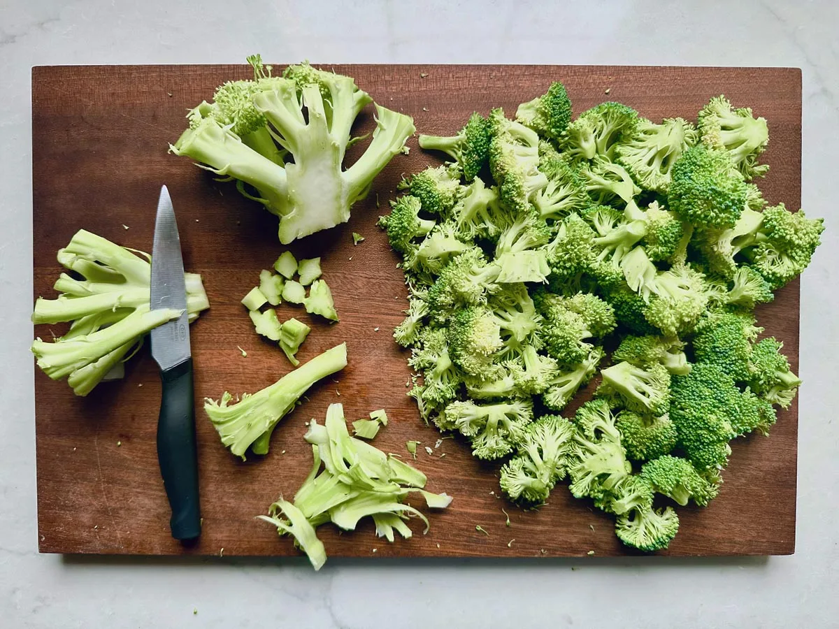 A wooden cutting board with a knife, chopped broccoli florets and stems, sliced carrots, and cubes of PepperJack cheese arranged on top, set on a light-colored surface.