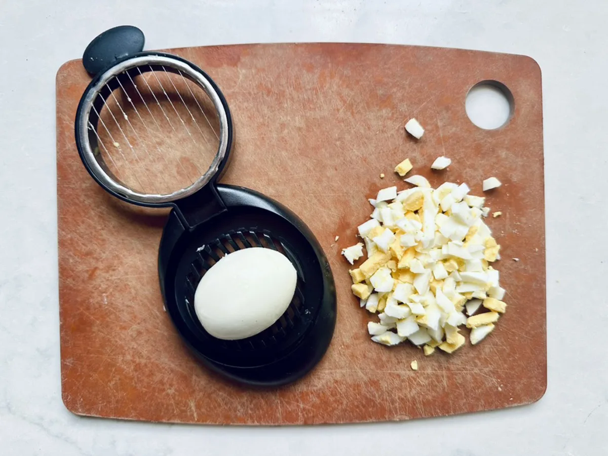 A hard-boiled egg sits in a black egg slicer on a brown cutting board. Next to it, chopped pieces of another hard-boiled egg are ready&mdash;perfect for making classic All-American Russet Potato Salad.