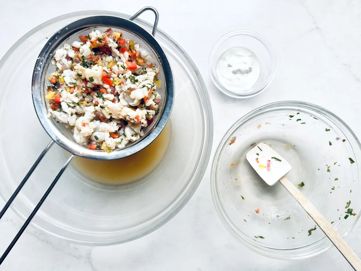 A strainer with chopped vegetables and herbs rests over a glass bowl containing liquid. Nearby, there is an empty glass bowl with a spatula and a small bowl with a creamy white sauce on a white surface.