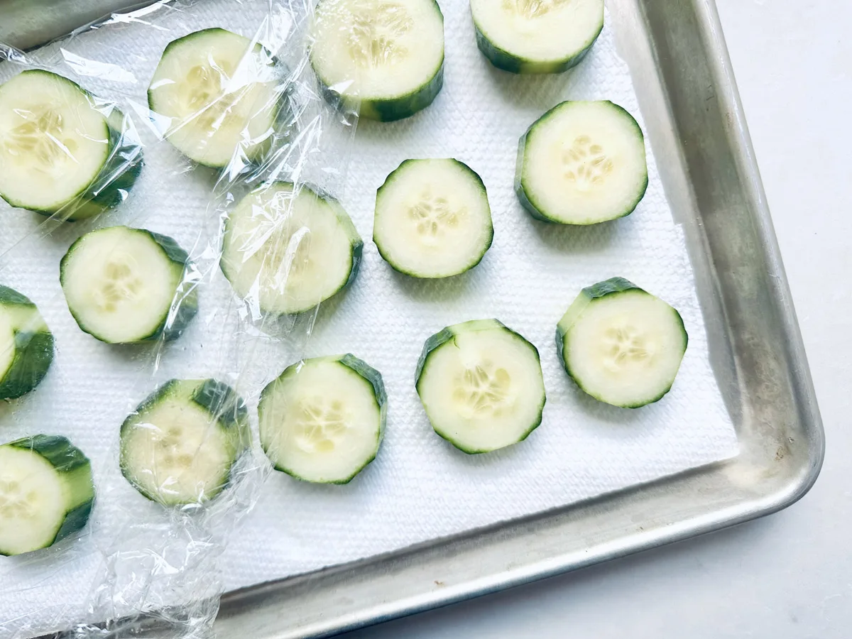 Sliced cucumbers arranged on a paper towel in a metal tray, partially covered with plastic wrap.