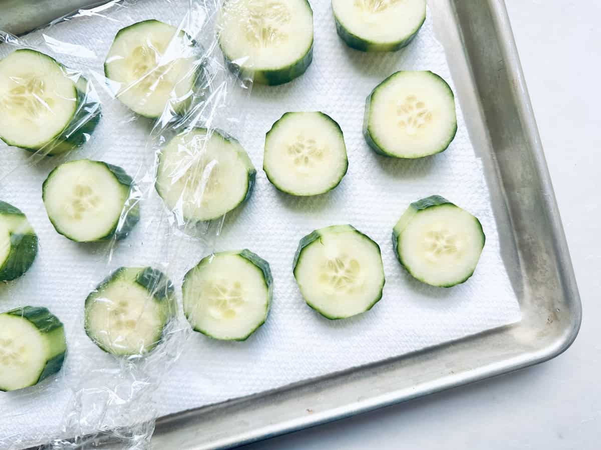 Sliced cucumbers arranged on a paper towel in a metal tray, partially covered with plastic wrap.