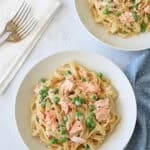 Two white bowls filled with fettuccine pasta, green peas, and flaked salmon in a creamy sauce are on a white marble surface, with a folded napkin, two forks, and a blue polka dot cloth nearby.