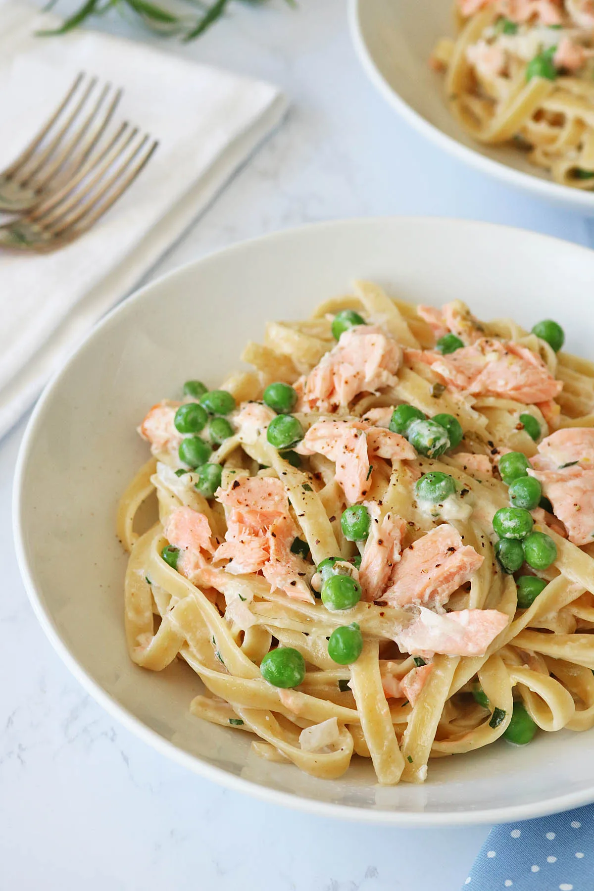 A white bowl filled with fettuccine pasta, green peas, and flaky salmon pieces in a creamy sauce. A napkin and forks are visible in the background on a white surface.