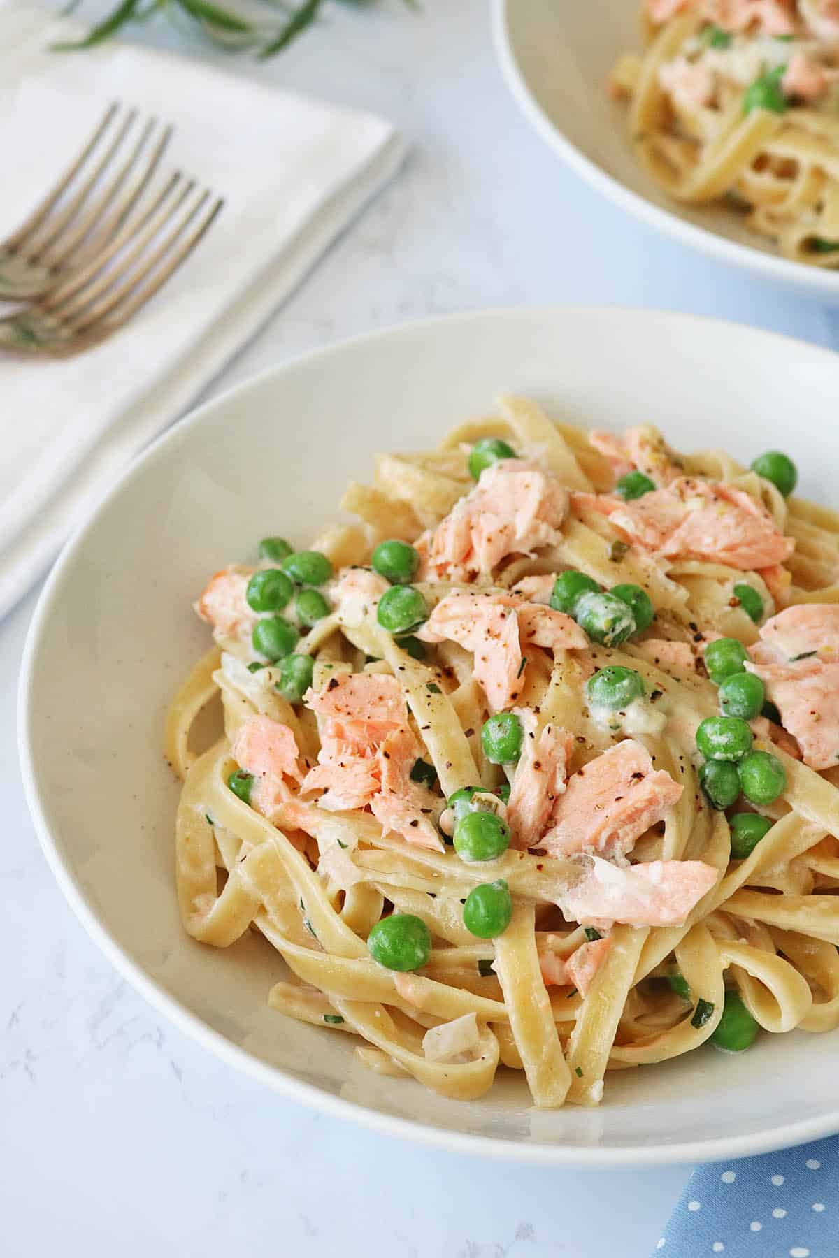 A white bowl filled with fettuccine pasta, green peas, and flaky salmon pieces in a creamy sauce. A napkin and forks are visible in the background on a white surface.