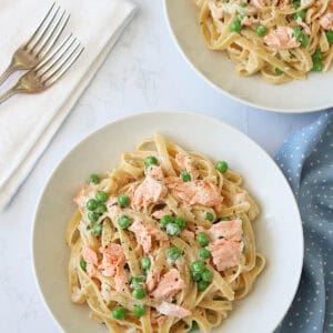 Two white bowls filled with fettuccine pasta, green peas, and flaked salmon in a creamy sauce are on a white marble surface, with a folded napkin, two forks, and a blue polka dot cloth nearby.