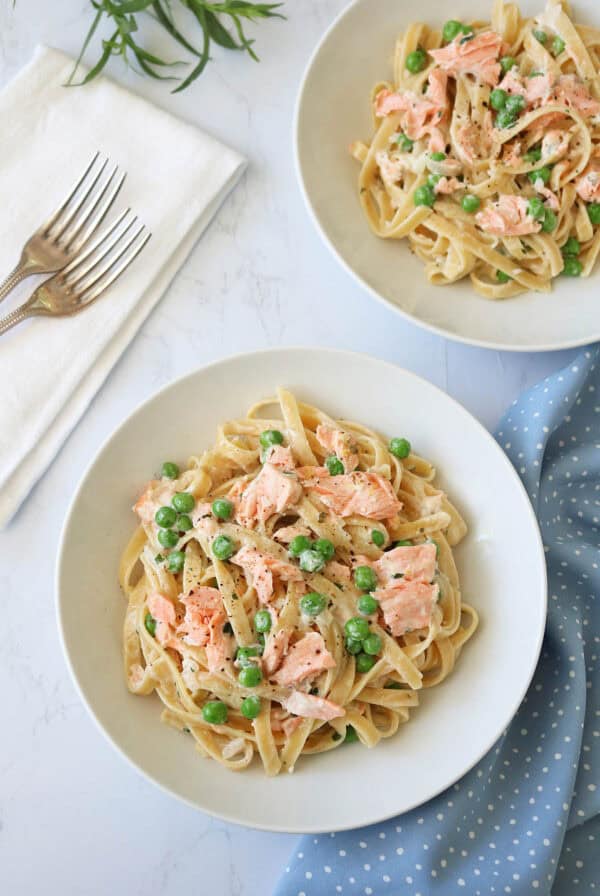 Two white bowls filled with fettuccine pasta, green peas, and flaked salmon in a creamy sauce are on a white marble surface, with a folded napkin, two forks, and a blue polka dot cloth nearby.