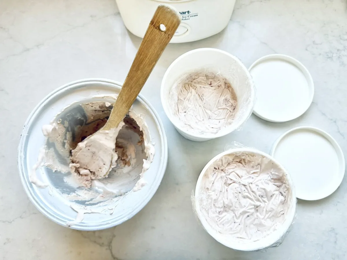 A mixing bowl with a wooden spoon and two containers filled with light pink Strawberry-Balsamic Ice Cream sit on a white countertop. The containers have lids nearby, suggesting homemade ice cream preparation.