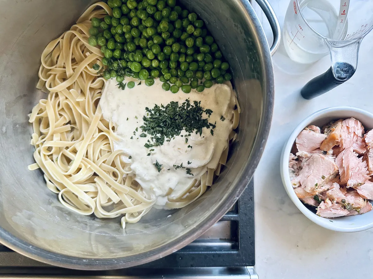 A metal bowl contains cooked linguine, green peas, a creamy white sauce topped with herbs. Next to it is a small bowl of flaked cooked salmon and a glass measuring cup with liquid on a countertop.