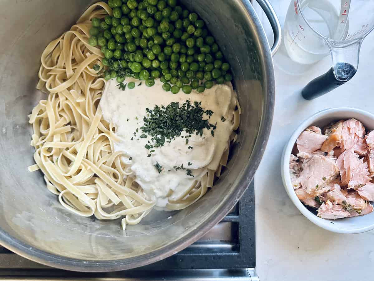 A metal bowl contains cooked linguine, green peas, a creamy white sauce topped with herbs. Next to it is a small bowl of flaked cooked salmon and a glass measuring cup with liquid on a countertop.