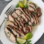Sliced grilled pork garnished with chopped herbs and served with lime wedges and cilantro on a white plate, with a fork and a small jug of sauce on the side.
