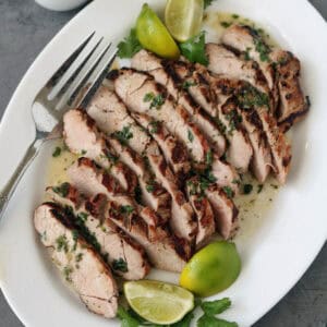 Sliced grilled pork garnished with chopped herbs and served with lime wedges and cilantro on a white plate, with a fork and a small jug of sauce on the side.