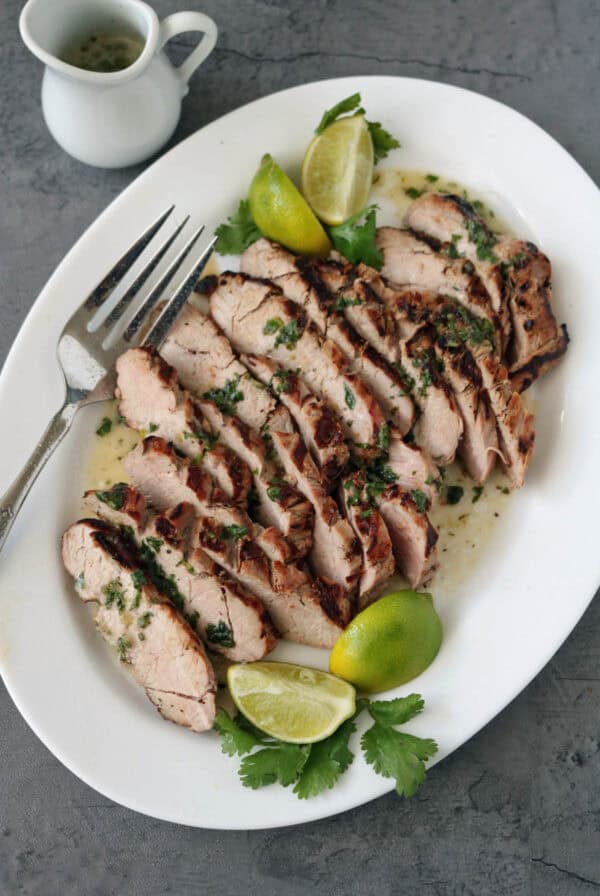Sliced grilled pork garnished with chopped herbs and served with lime wedges and cilantro on a white plate, with a fork and a small jug of sauce on the side.