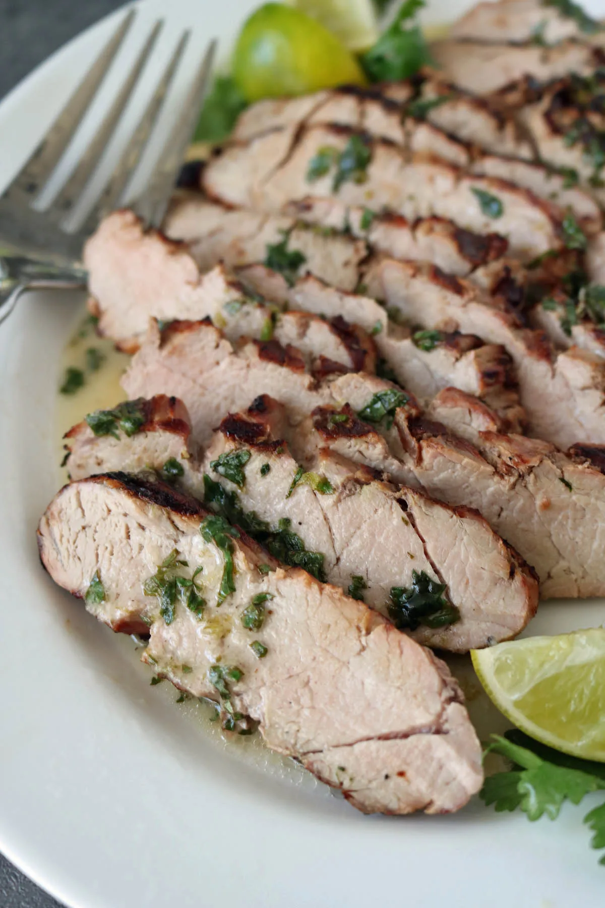 Sliced grilled pork garnished with fresh herbs and lime wedges, served on a white plate with a serving fork.