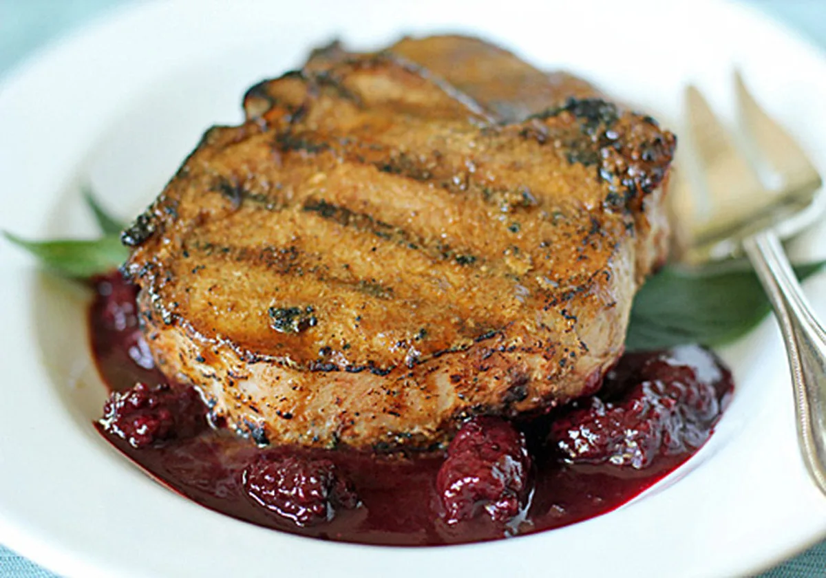 A grilled pork chop sits on a white plate over a bed of dark red berry sauce, garnished with green leaves, with a fork and knife placed next to it.