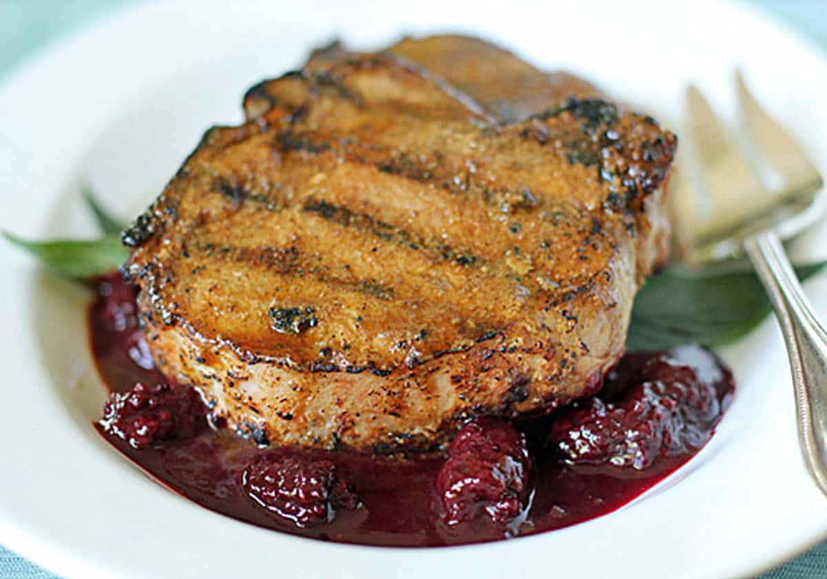 A grilled pork chop sits on a white plate over a bed of dark red berry sauce, garnished with green leaves, with a fork and knife placed next to it.