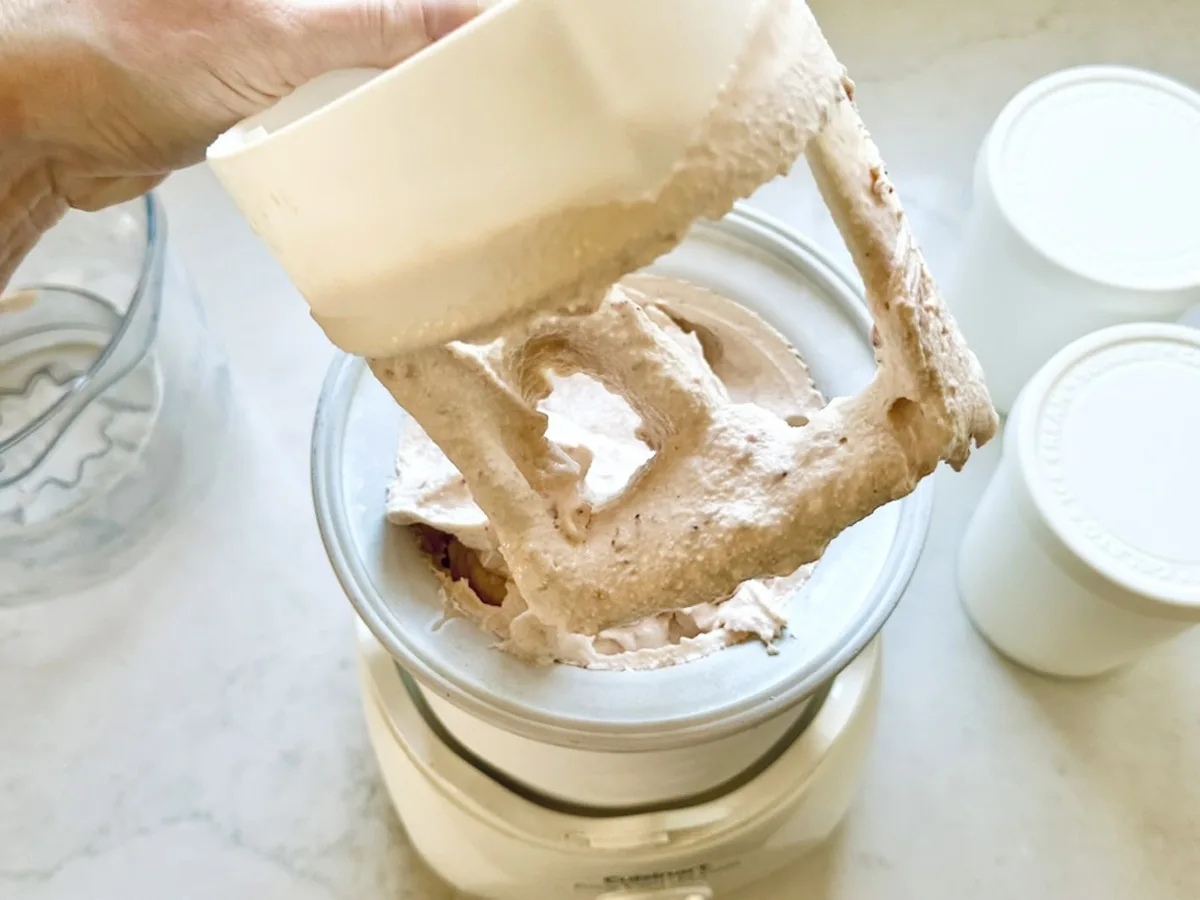 A hand lifts a mixer attachment coated with light brown, whipped Strawberry-Balsamic Ice Cream from an ice cream maker, with two white containers nearby on a countertop.