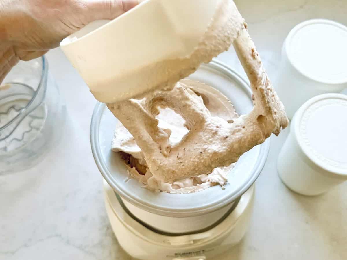A hand lifts a mixer attachment coated with light brown, whipped Strawberry-Balsamic Ice Cream from an ice cream maker, with two white containers nearby on a countertop.