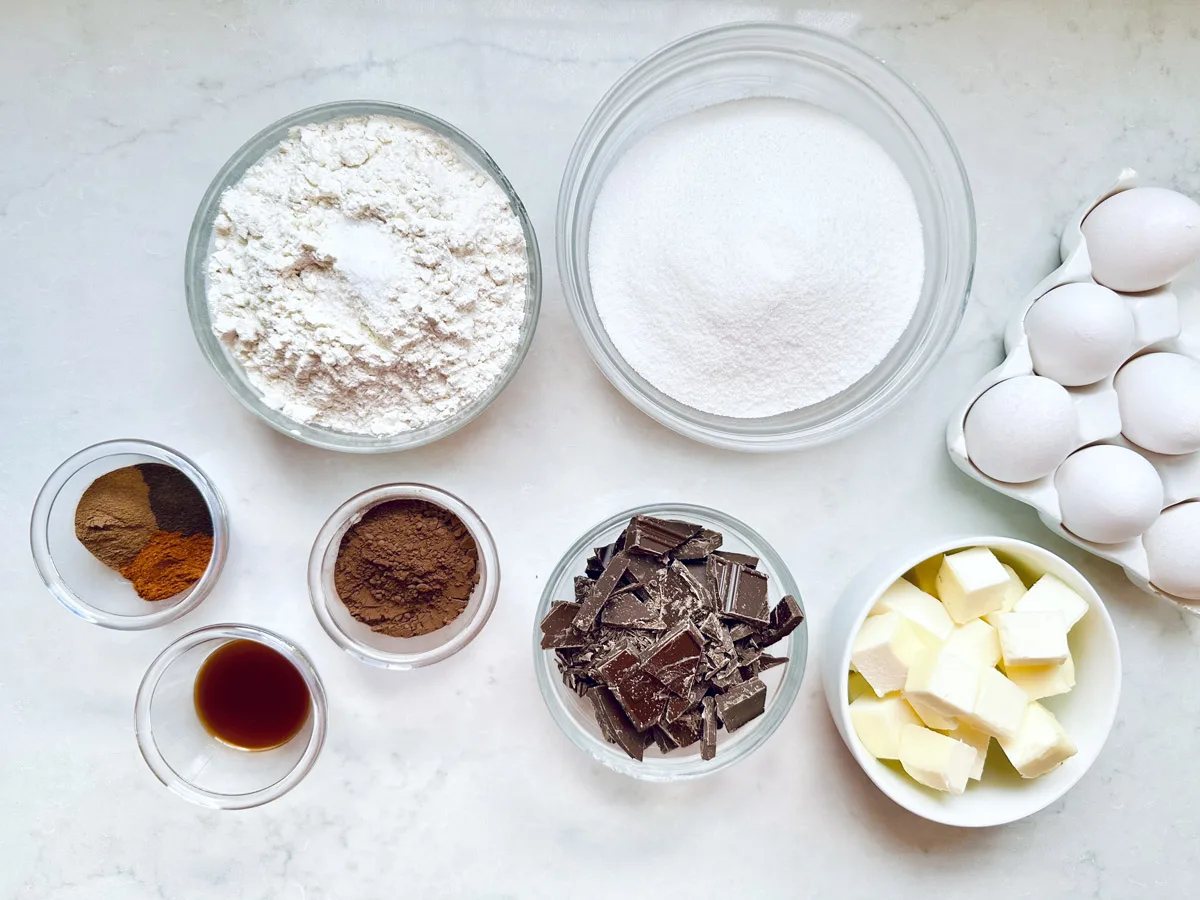 Assorted baking ingredients on a white surface: bowls of flour, sugar, chocolate chunks, cocoa powder, cinnamon, butter cubes, vanilla extract, and a carton of eggs—perfect for making Chocolate Chipotle Brownies.