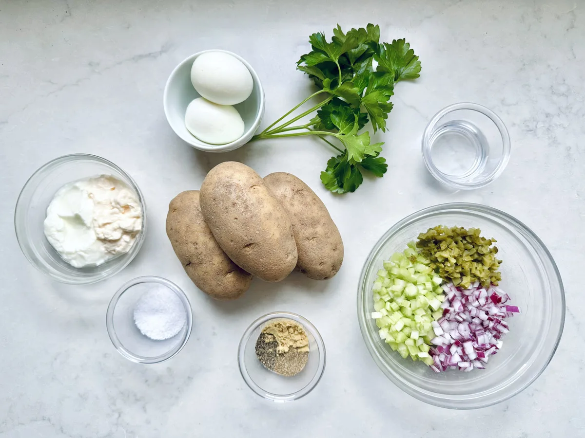 Ingredients for All-American Russet Potato Salad arranged on a white surface, including potatoes, hard-boiled eggs, parsley, chopped celery, pickles, red onion, mayonnaise, mustard, salt, pepper, and water in small bowls.