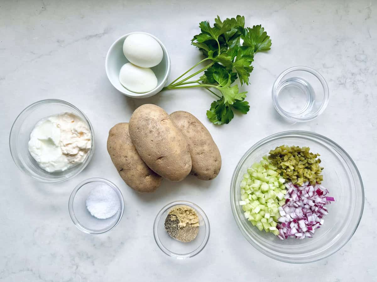Ingredients for All-American Russet Potato Salad arranged on a white surface, including potatoes, hard-boiled eggs, parsley, chopped celery, pickles, red onion, mayonnaise, mustard, salt, pepper, and water in small bowls.