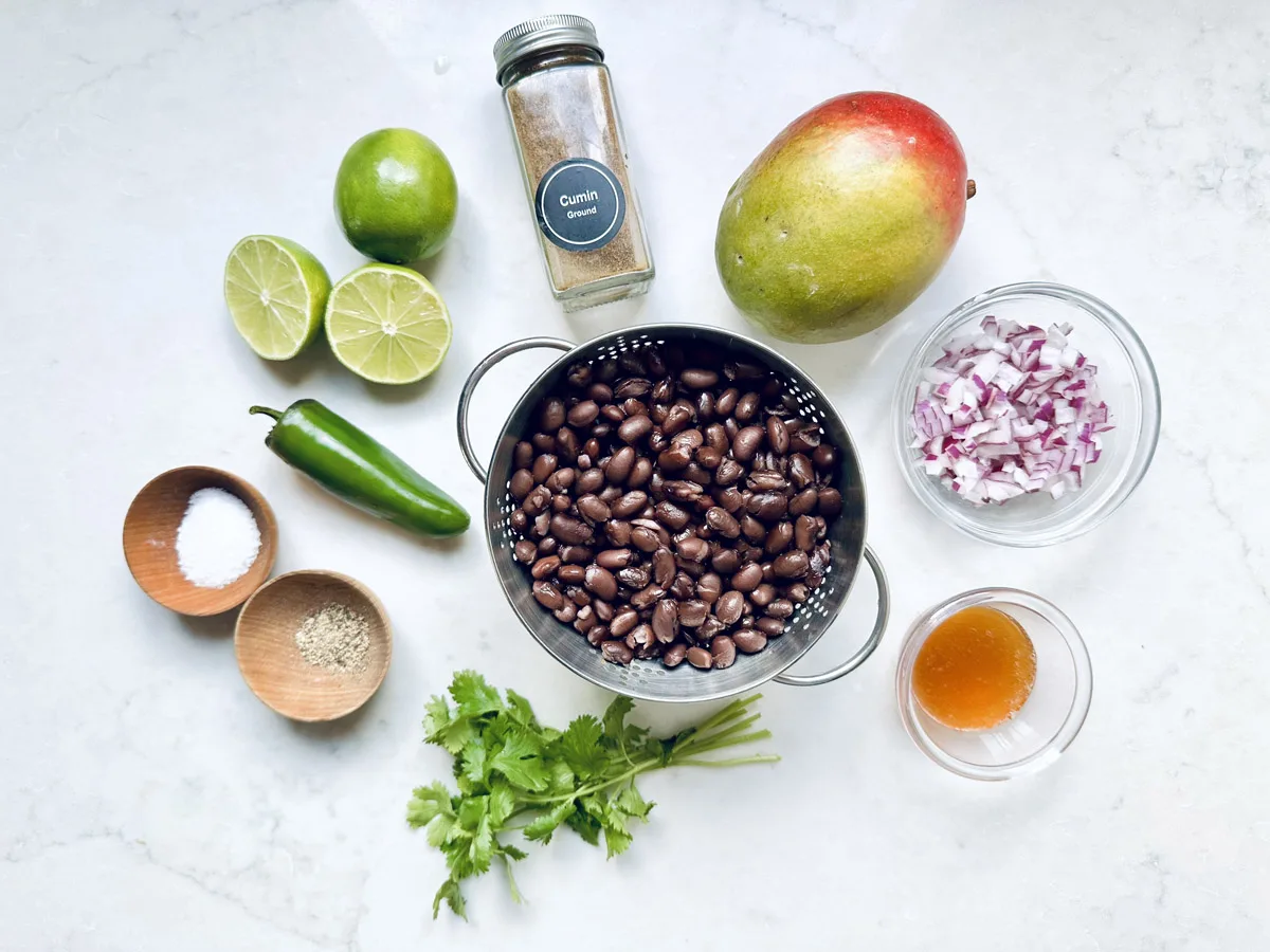 Ingredients for a black bean salad arranged on a white surface: black beans in a colander, diced red onion, mango, lime halves, cilantro, sliced jalapeño, cumin, salt, pepper, and a small bowl of honey.