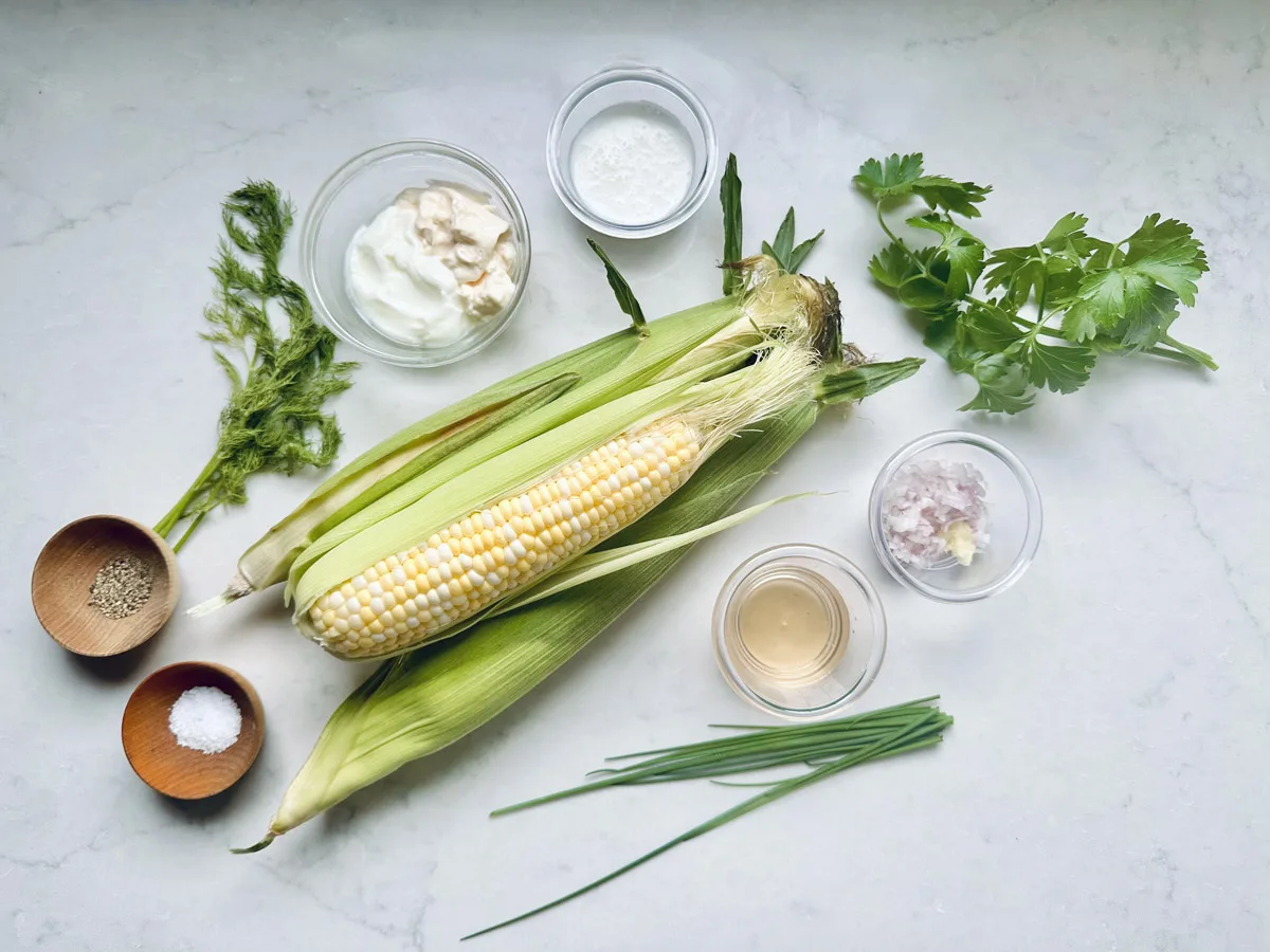 Fresh corn in husks, fresh herbs, yogurt, vinegar, minced garlic and shallot, and small bowls of salt and pepper are arranged on a white surface—perfect for preparing Corn Salad with Buttermilk and Herbs.