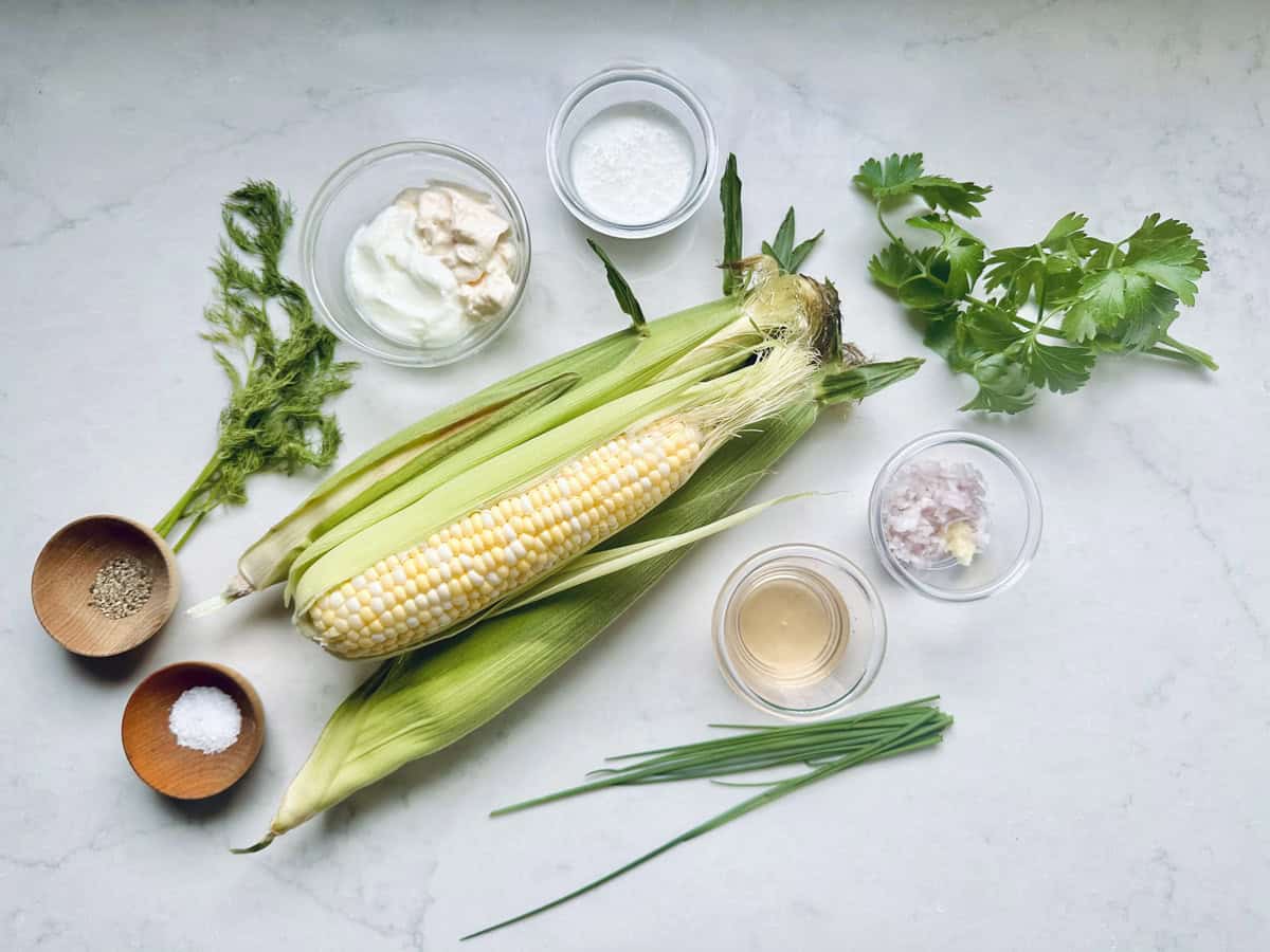 Fresh corn in husks, fresh herbs, yogurt, vinegar, minced garlic and shallot, and small bowls of salt and pepper are arranged on a white surface—perfect for preparing Corn Salad with Buttermilk and Herbs.