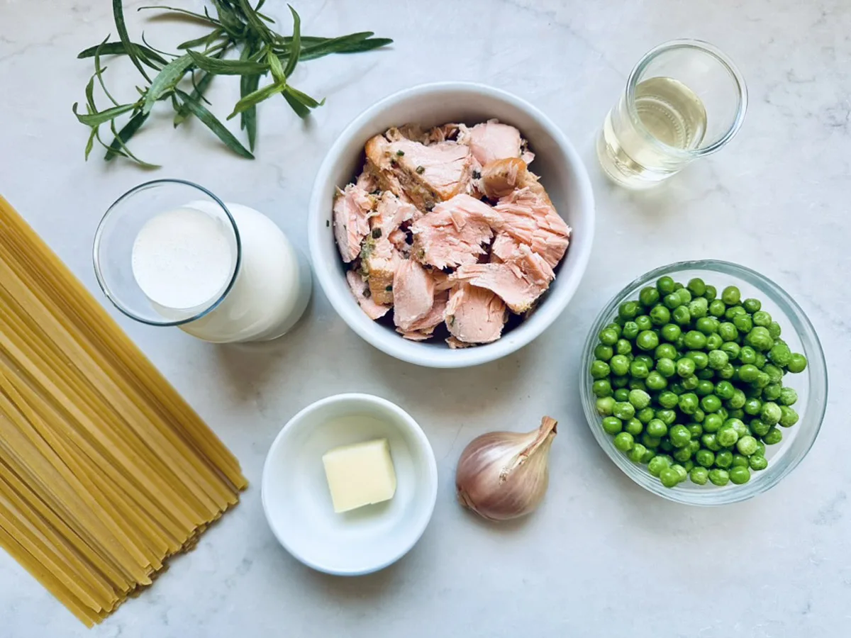 Uncooked spaghetti, fresh tarragon, a glass of milk, a small glass of white wine, a bowl of cooked salmon chunks, a bowl of green peas, a shallot, and a pat of butter arranged on a white surface.