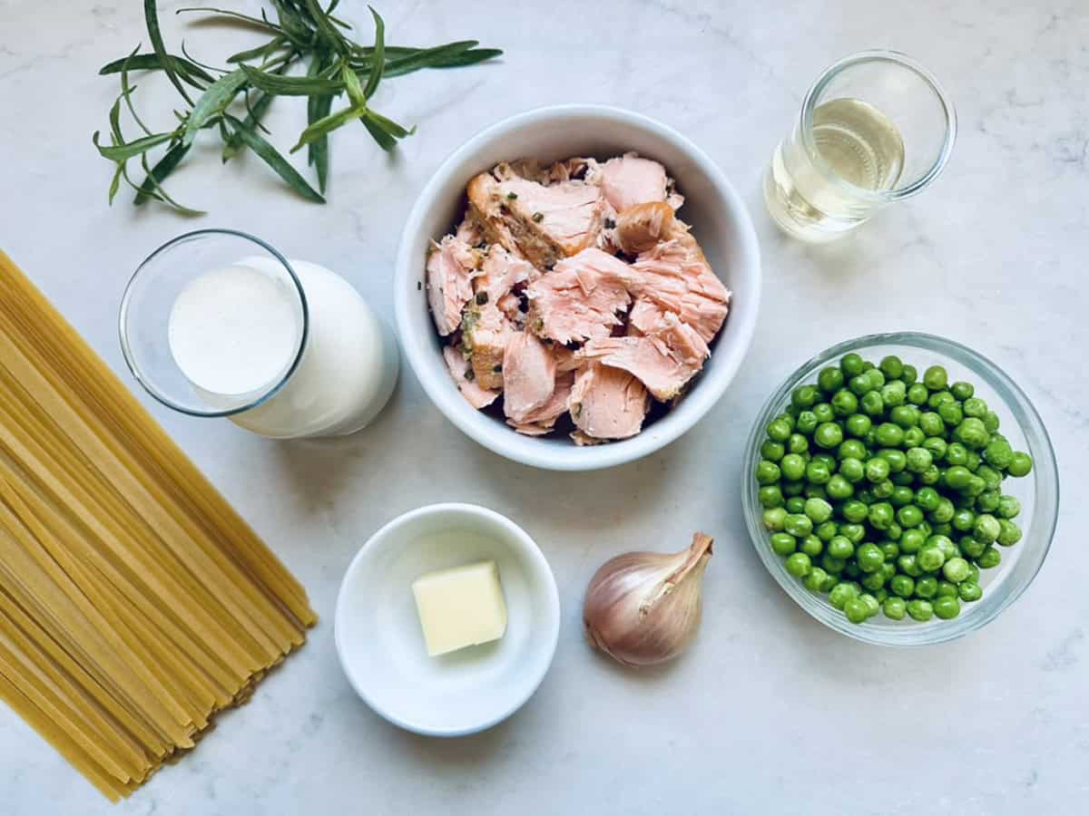 Uncooked spaghetti, fresh tarragon, a glass of milk, a small glass of white wine, a bowl of cooked salmon chunks, a bowl of green peas, a shallot, and a pat of butter arranged on a white surface.